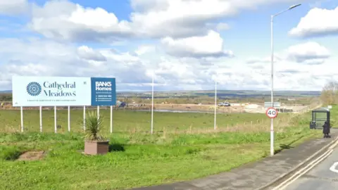 The site of the development, photographed from Station Road in West Rainton. It is a large grass land with a blue sign reading Cathedral Meadows Banks Homes. There is a 40mph speed restriction sign. A person is waiting near a bus shelter on the side of the road. 