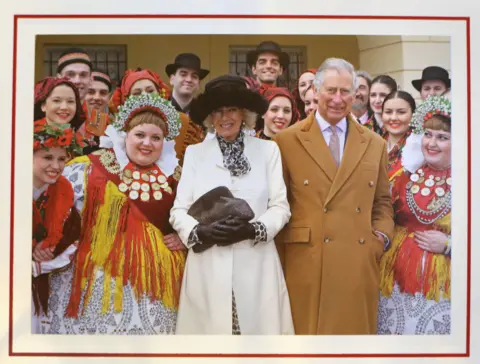 PA Media Prince Charles and Camilla's 2016 card, showing the couple during a visit to Croatia, standing in winter coats - Camilla with warm hat and gloves, Charles with his hands in his pockets. Performers dressed in costumes are standing either side of the royal couple and behind them.