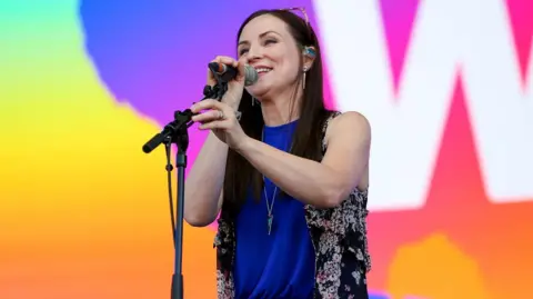 Julie Fowlis is standing on a stage at a BBC Big Weekend event. She has long dark hair, and has a pair of sunglasses on her head. She is singing into a microphone.
