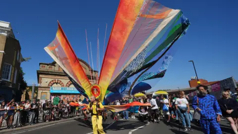PA Media A performer in an all yellow outfits wears a huge rigged costume with rainbow sails pointed into the air.