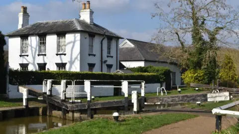 Andrew Abbot/Geograph Leighton Lock Cottage, a two-storey white buidling, with Leighton Pumping Station, another white building, behind. Both are on the opposite side of a canal lock