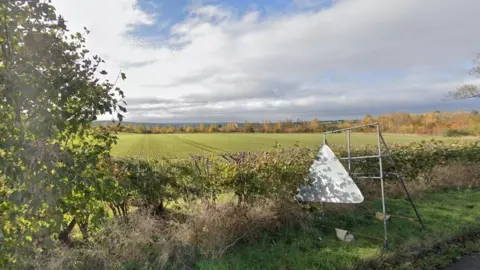 A large green field framed by a green tree and hedgerows under a cloudy sky. To the right, the back of a triangular sign resting on a trestle.
