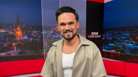 A head and shoulders image of a man smiling at the camera in a TV news studio. He has short dark hair, a trimmed beard and is wearing a white top with a beige unzipped collared shirt over the top. 