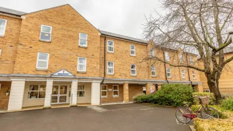 St George's Court care home is a brick building with three storeys. There is a large entrance door and a number of windows. In front is a parking area and a bicycle can be seen parked near bushes and a tree.