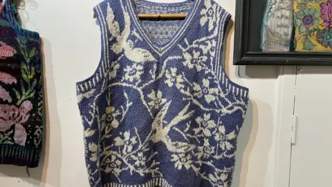 BBC, Leanne Rinne A blue and white knitted vest jumper hanging on a wall next to a painting. The pattern has birds on it and flowers. 