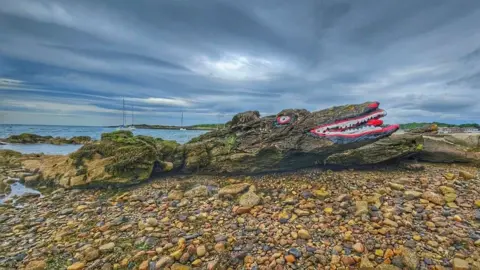 The face of a crocodile painted onto a long rock on a pebble beach