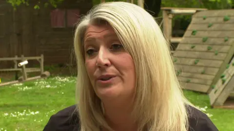 A woman with blonde hair speaking at the camera. It is a head shot and she is outside. There is green grass behind her and a wooden children's play area.