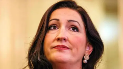 PA Media Emma Little Pengelly looking straight ahead. She has long brown hair and is wearing gold earrings with a pearl hanging from them.