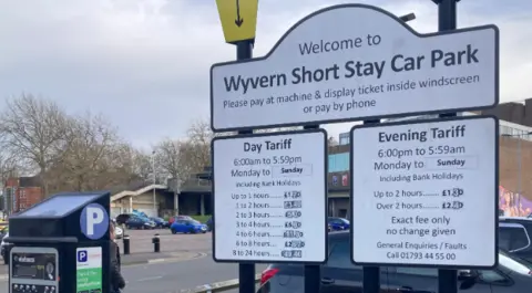 Car park ticket machine and white tariff boards in the foreground with parked cars in the background