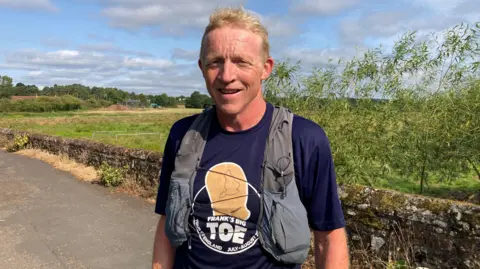 BBC Frank Wainwright smiling at the camera standing on a lane with a field behind him
