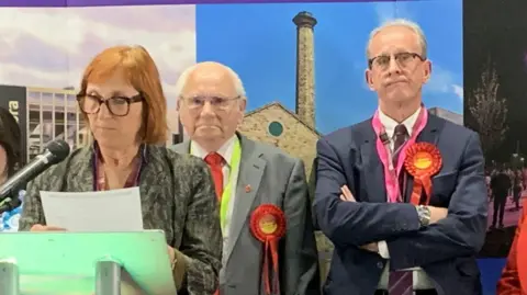 BBC/Lucy Ashton A woman is stood at a podium reading from a piece of paper, she has auburn hair in a bob, glasses and a grey blazer. Behind her on the right is a man with a balding head, white hair and glasses wearing a grey suit and a red Labour rosette. On the left is a man with grey hair and glasses looking fed up and annoyed in a dark suit with a red Labour rosette