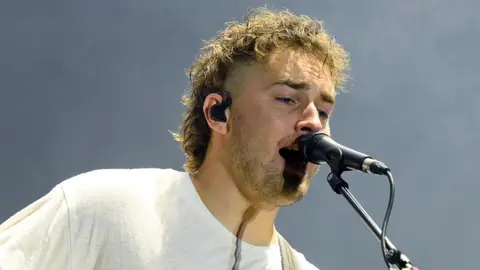 Getty Images Sam Fender wearing a white tshirt singing into a black microphone with a black ear piece in his right ear.