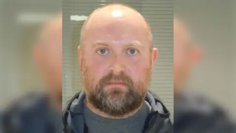 Lincolnshire Police Police custody photograph of Karl Abbott who is bald with a dark beard and wearing a grey T-Shirt and jacket