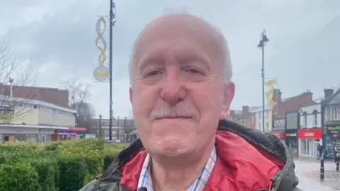 A bald man with a moustache is stood in a town centre shopping street. The lampposts behind him have Christmas decorations on them and there is a row of planted bushes to the left hand side of the image.