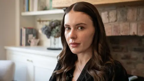 A dark haired woman in a black top looks sideways at the camera. In the background in a chimney breast and a bookshelf