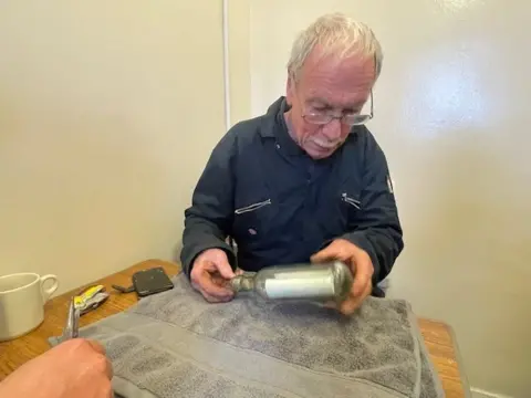 Ross Russell Barry is examining the bottle. He is sitting at a table and has white hair and is wearing glasses and a blue work suit.