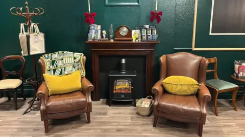 A bookshop room with wooden flooring and a dark green wall. There are two brown leather armchairs either side of a small electric fireplace with a wooden mantelpiece above it. The chairs have blankets and yellow cushions on them