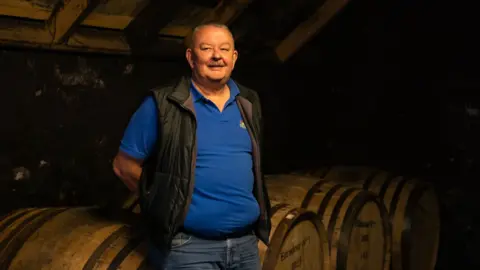 Edradour Distillery Andrew Symington has grey hair and smiles at the camera with his hands behind his back while standing in a dimly-lit room in front of whisky casks lying on their side. He wears a blue polo shirt, a black gillet and blue jeans.