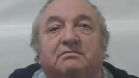 A man with thinning grey hair in a close-up police custody image. He is standing in front of a grey background. 