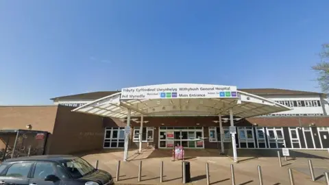 Google The outside of the front of a hospital. It is a brown brick building with a white canopy at the front and white framed windows. In front of the building are several bollards, and a black car parked on the road.