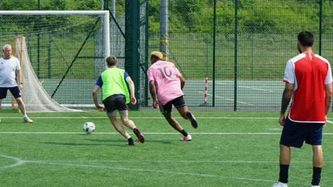 A woman in a luminous yellow sports bid and another man in a pink football jersey race towards a football. A man in a white t-shirt and black shorts guards a white goal post. They are playing on green artificial grass lined with white football field markings. The pitch is surrounded by a dark green mental fence.