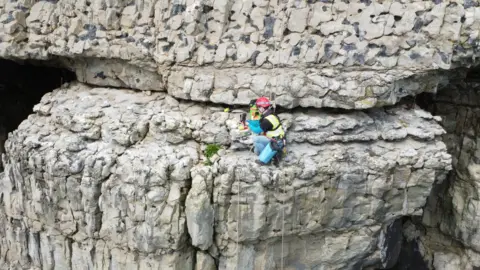 James Preston A climber on a ledge sheer rock face, with ropes handing down, working with camera equipment.