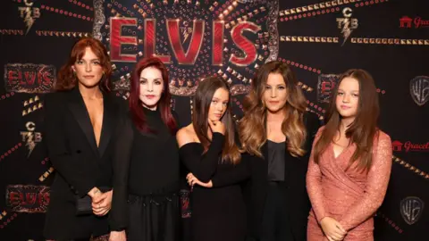 Getty Images for Warner Bros. Riley Keough, Priscilla Presley, Harper Vivienne Ann Lockwood, Lisa Marie Presley and Finley Aaron Love Lockwood at a special screening of ELVIS on 11 June 2022. They are all wearing black clothes, except Finley who is wearing a peachy-pink lace dress. They are standing in front of the advertising poster for the Elvis movie.