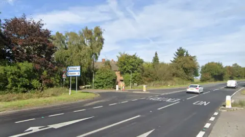 Two-lane dual carriageway, with a sign off to Tiffield and Gayton to the right. There are lane markings indicating that the left lane is for proceeding straight ahead or turning towards Tiffield. There is a bollard to the right indicating that there is a central reservation for vehicles heading south. The road is lined with trees.