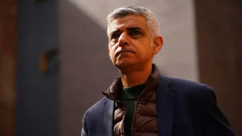 Mayor Sadiq Khan wearing a copper brown puffer gilet and dark green jumper under a navy woollen coat. He has short white and grey hair and is looking slightly upwards. The background is blurred.