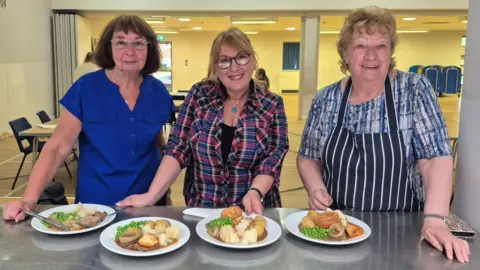 Bournville Village Trust Three women are standing next to a metal table with plates of potatoes, peas, carrots and gravy. They are standing in a village hall with tables and chairs laid out.