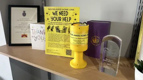 A shelf which has a certificate, a thank you card and an engraved glass Queen's Jubilee Award on it. There is also a yellow charity collection box, with a yellow poster behind it titled 'we need your help'.