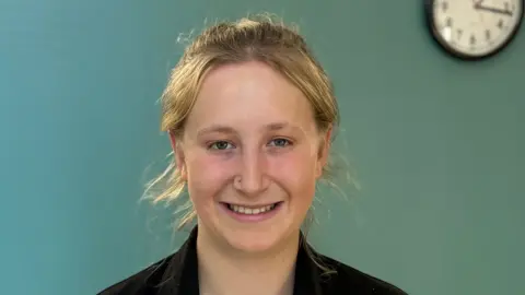 HTP Apprenticeship College Annabelle smiling, standing in front of a blue wall with a clock in the background