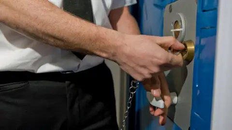 Close-up shot of a prison officer opening a cell door. The officer is wearing a white shirt and black trousers and the key is attached to a chain. The brass-coloured lock is set in a grey panel on a blue door.