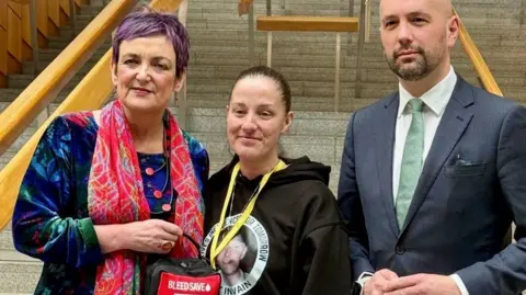 Three individuals standing together inside a building with wooden handrails and wide steps in the background. One person is wearing a brightly patterned outfit and holding a small red-and-black bag labelled ‘BLEEDSAFE.’ The person in the middle is wearing a dark hoodie with a circular graphic on the front. The person on the right is dressed in a suit with a light-colored shirt and patterned tie.”
