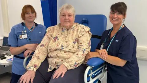 Derbyshire Community Health Services A woman with white hair and patterned shirt sits in a blue hospital chair. Either side of her are two health workers.