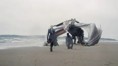 Theo Whiteman and HBO Scene from House of the Dragon - two actors on a beach, one kneeling, with a dragon behind him