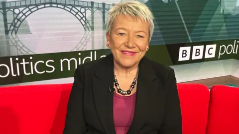 Rachel Taylor - a woman with short blonde hair and wearing a black suit jacket over a purple top, smiles as she sits on the Politics Midlands red sofa.