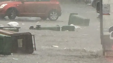 Bins have fallen into flood water on a road in Plymouth. A red car is travelling past the floating bins. Heavy rain is pouring down.