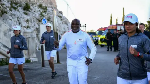 CHRISTOPHE SIMON/AFP French former football player Basile Boli holds the Olympic Torch as part of the Olympic and Paralympic Torch Relays at the Notre Dame de la Garde Basilica