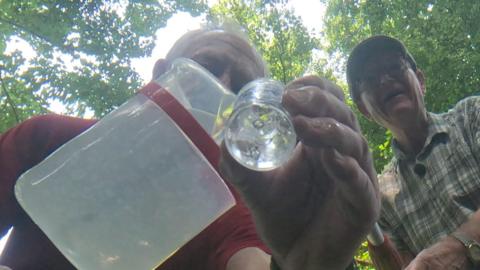 Two men shot from below one pouring river water into a glass jar with a leafy tree canopy behind them