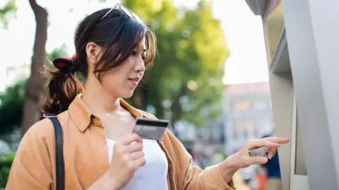 A woman uses a cash machine on the street on a sunny spring day. She holds a credit card in her hand and is pressing buttons on the machine with her other hand. 
