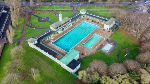 BBC A drone image of the Peterborough Lido. The site features a large and small swimming pool with shrubs, trees and paving around the perimeter. 