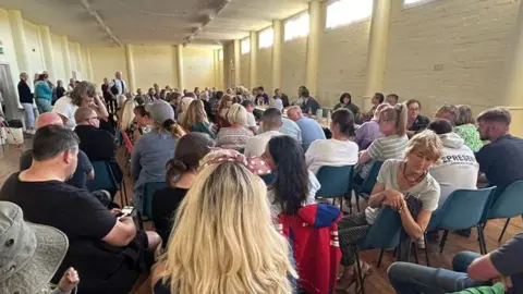 About 100 men and women sit on chairs in a yellow hall to listen to a panel of speakers.