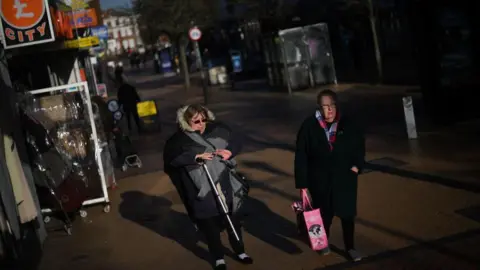 Getty Images Women walk down the high street in Grays on January 18, 2023. They are wearing black coats and one is holding a walking stick.