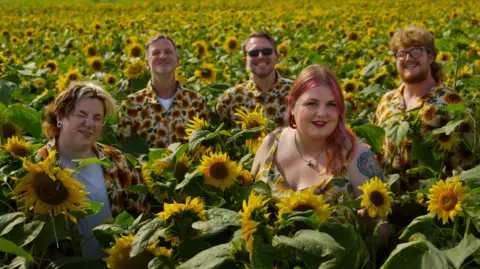 Mark Mangan The five-piece band stand in a field of sunflowers. They are all wearing tops with sunflowers on them and smiling at the camera.