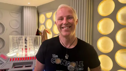 A man with short blond hair is smiling for a head and shoulders photo and is wearing a black top that says The Console Room on it. He is also wearing a circular necklace on a black thin rope. Behind him are grey walls with yellow circles on it, a hat stand with a hat and brown coat and a scarf on the other side. Next to him is a clear cube on a console with several buttons.