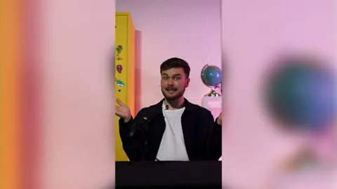 Conor, with short brown hair, a moustache and short beard, mid-speaking, wearing a white t-shirt and black jacket. The background features a white wall a yellow locker to the left side and a world globe on the right. 