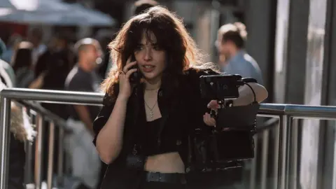 We Stream A woman with tousled, shoulder length black hair wearing a leather jacket over a tube top chats on a mobile phone as she holds a large, professional looking camera in the other hand. She's leaning against a metal railing, and a small crowd mills about behind her.