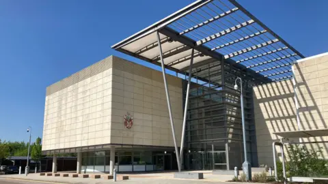 Emma Howgego/BBC South Cambs council building which is a modern blocky structure made of beige blocks and a recessed glass section in the middle. There is a large dark grey metal pergola structure held up by V-shaped poles. Clear blue sky behind.