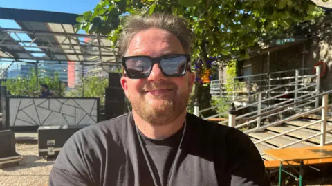 The DJ Eats Everything in a black t-shirt and big black sunglasses, smiling. He is standing in the back yard of Motion, with a staircases and a small stage behind him and some flight cases on the floor.